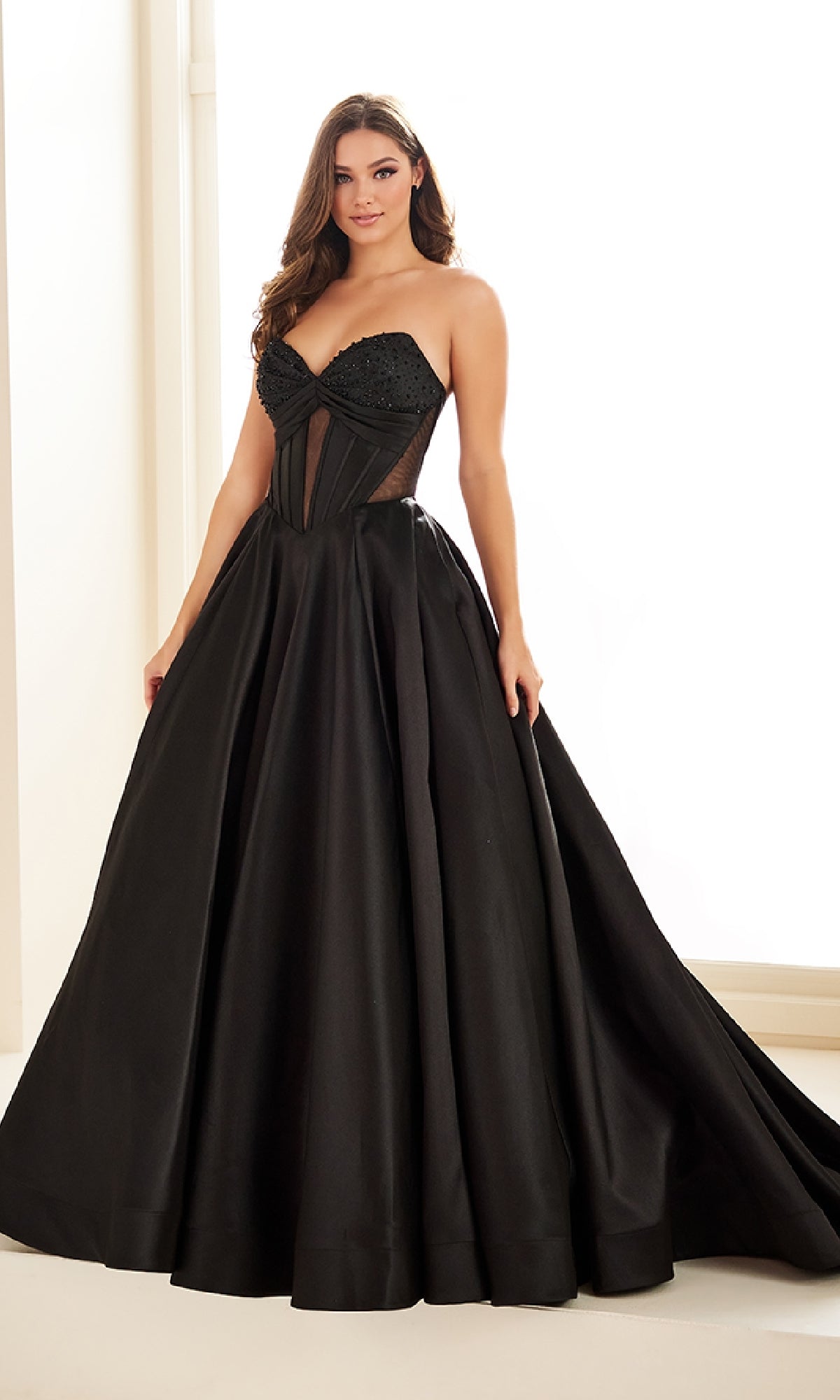 A woman stands indoors wearing the Ellie Wilde EW36025, a strapless black satin prom ball gown with a fitted corset bodice, dramatic full skirt, and an elegant lace-up open back for added sophistication.